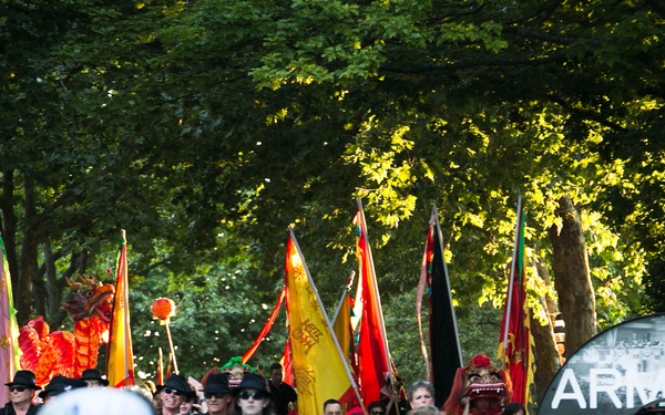 A festive outdoor parade is taking place near a building marked 'ARMORY,' featuring a marching band in blue and yellow uniforms, flag bearers carrying colorful flags, and what appears to be a Chinese dragon costume. The event draws a large crowd of spectators and participants along a tree-lined path. The scene suggests a multicultural celebration or festival.