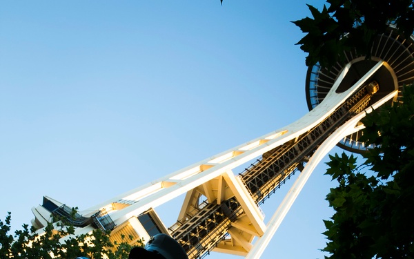 A low-angle upward shot captures uniformed military personnel, identifiable as U.S. Marines based on their dress uniforms and rank insignia, standing beneath Seattle's Space Needle. The dramatic perspective emphasizes the tower soaring into a clear blue sky, with trees framing the composition. The warm lighting on the Space Needle suggests the photo was taken during golden hour (early evening or late afternoon).