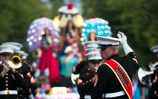 A United States Marine Corps marching band performs in a parade, led by a drum major in formal dress uniform with a red sash and white gloves. Behind the band, illuminated parade floats are visible, including one featuring women in formal gowns who appear to be pageant contestants. The scene takes place on a public street lined with spectators and trees.