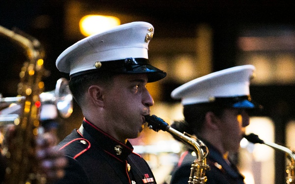 A group of U.S. Marine Corps band members are performing outdoors at night, playing saxophones in full dress uniform. The central figure is sharply in focus, actively playing an alto saxophone while wearing the distinctive Marine Corps dress blues with white cover (hat). Multiple musicians are visible in the background, slightly out of focus.