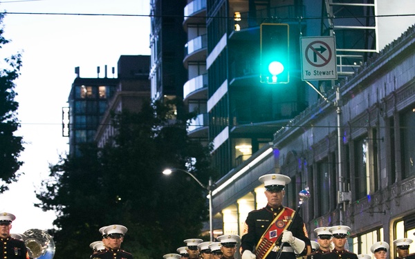 A U.S. Marine Corps marching band performs in a nighttime street parade in an urban setting. The band members are dressed in formal dress blues uniforms with white covers, led by a drum major carrying a ceremonial mace. Spectators line the sidewalks behind barriers to watch the procession.