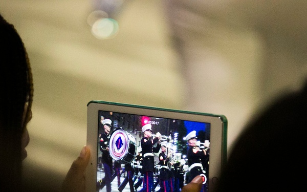 A person is holding a tablet device and recording or viewing a military marching band parade. The tablet screen displays uniformed band members marching with instruments, including a large bass drum bearing a military insignia. The foreground shows the silhouetted heads of spectators in a darkened environment.