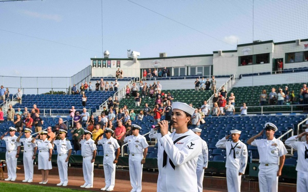 A U.S. Navy sailor in dress white uniform performs what appears to be the National Anthem at a minor league baseball stadium, holding a microphone while fellow sailors stand at attention or salute in a line behind them. The ceremony is taking place on the field before a game, with a crowd of spectators filling the stadium stands. The setting and uniforms suggest a military appreciation night or similar patriotic pre-game ceremony.
