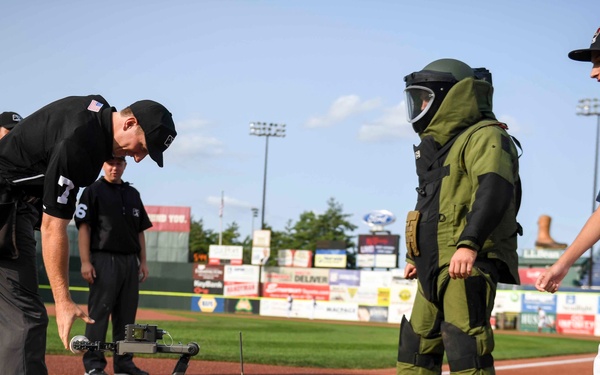 A pre-game or promotional event at a minor league baseball stadium features a bomb disposal robot (EOD robot) near home plate. An umpire in black uniform bends down to interact with the tracked robot, while a person in a full bomb disposal suit stands nearby alongside a young baseball player wearing jersey number 88. The scene appears to be a special demonstration or fan engagement event at the ballpark.