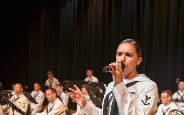 A U.S. Navy enlisted sailor in dress white uniform performs as a vocalist at a formal military band concert on a wooden stage. Behind her, a large ensemble of Navy musicians in matching dress whites are seated with various instruments including brass, woodwinds, and percussion. The setting appears to be an indoor auditorium or performance hall with dark curtains in the background.
