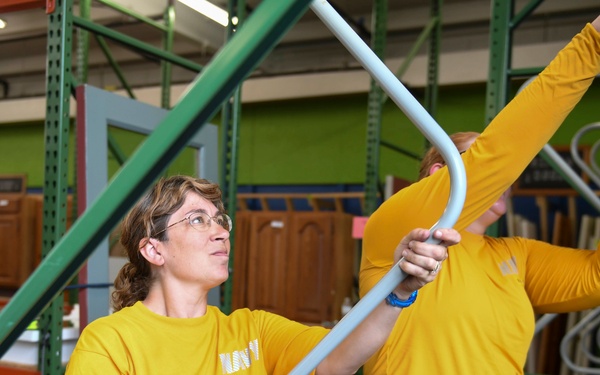 Two individuals wearing matching yellow t-shirts are working together in what appears to be a warehouse or resale store. They are collaborating to handle a large curved metal or PVC pipe/frame, with one person holding the lower end and the other securing the upper end with a tool. The background shows green metal shelving racks and various furniture items in storage.