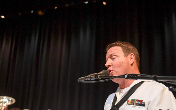 A U.S. Navy Petty Officer in dress white uniform is playing a keyboard synthesizer and singing into a microphone on a performance stage. Behind him, additional Navy personnel in white uniforms are playing brass instruments and drums. The setting appears to be a formal military band concert performance.