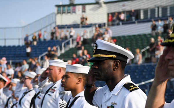 A line of U.S. Navy personnel in white dress uniforms are standing at attention and saluting during a ceremony at a baseball stadium. The formation includes both enlisted sailors and officers of varying ranks, identifiable by their insignia, ribbons, and cap designs. Spectators are visible in the stadium stands in the background.