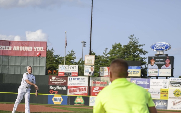 A person in white military dress uniform is throwing a ceremonial first pitch at a minor league baseball stadium. A catcher in a neon yellow shirt is crouched at home plate ready to receive the throw. The stadium outfield wall is lined with numerous sponsor advertisements.