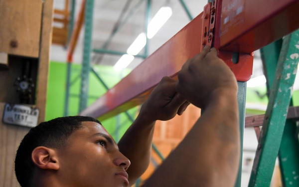 A young man in a yellow t-shirt is working on assembling or adjusting industrial metal shelving/racking in what appears to be a warehouse or storage facility. He is reaching upward with both hands to manipulate a red horizontal beam connecting to teal/green vertical rack uprights. The background shows a colorful interior space with fluorescent lighting and painted walls.