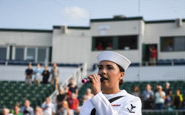 A U.S. Navy sailor in dress white uniform performs a vocal solo into a microphone at what appears to be a baseball stadium. Behind her, fellow Navy personnel in dress whites stand at attention and salute, suggesting a national anthem or ceremonial performance. The event appears to be a military appreciation day at a minor or major league baseball venue with spectators in the stands.