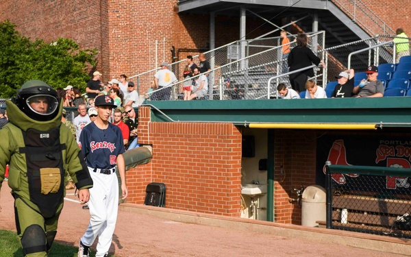 A bomb disposal technician in full EOD (Explosive Ordnance Disposal) suit walks alongside a baseball player wearing a Portland Sea Dogs uniform at what appears to be Hadlock Field in Portland, Maine. A tracked EOD robot is visible on the grass in the foreground. This appears to be a pre-game demonstration or public safety awareness event at a minor league baseball stadium.