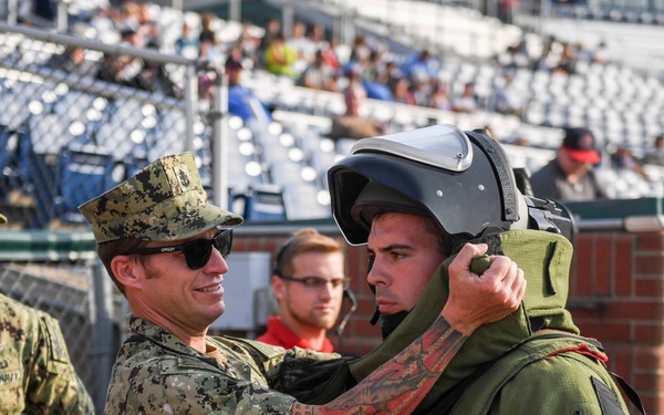 A U.S. Navy servicemember in digital camouflage uniform is helping a civilian try on an EOD (Explosive Ordnance Disposal) bomb suit, including a protective helmet, at what appears to be a public outreach or military appreciation event at a baseball stadium. The setting suggests a community engagement or military demonstration event with spectators visible in the stands. Several other Navy personnel and civilians are present in the background.