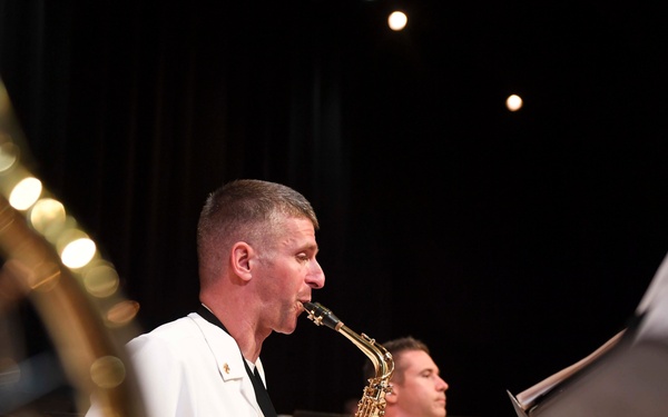 Military musicians in white dress uniforms perform in a concert band setting, playing saxophones on a darkened stage. The foreground subject is actively playing an alto saxophone while reading from a music stand. Multiple performers are visible in the background, all wearing matching white naval dress uniforms.