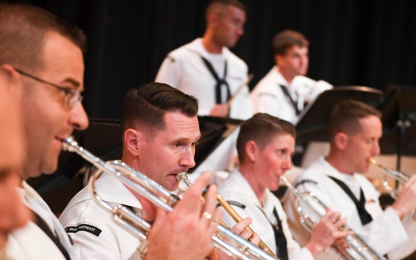 A military band, identifiable by their white dress uniforms with black neck ties, is performing a concert. Multiple musicians are seated in rows playing brass instruments, primarily trumpets, in what appears to be an indoor performance venue. A patch visible on one uniform reads 'NAVY NORTHEAST', identifying this as a U.S. Navy band.