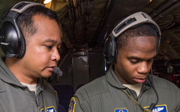 Two military personnel in olive green flight suits are inside what appears to be a military aircraft, both wearing aviation headsets. They are collaborating over a ruggedized tablet device, with one individual operating the touchscreen. Their uniforms display Air Force patches and name tapes, indicating they are U.S. Air Force aircrew members.