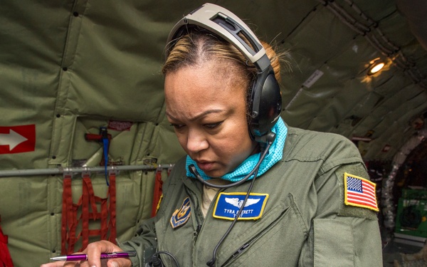 A female U.S. Air Force service member, identified by her name tag as 'Tyra Barrett', is seated inside a military aircraft working with a tablet device and paperwork. She is wearing an olive green flight suit with a U.S. flag patch, aviator wings insignia, and communication headset. The interior of the aircraft shows military cargo configuration with green padding, red safety straps, and emergency exit markings.