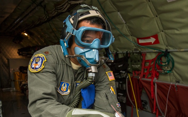 A U.S. Air Force personnel member wearing an oxygen/emergency breathing mask and flight suit is handling yellow oxygen or gas cylinders inside what appears to be a military transport aircraft. The individual is equipped with protective gear including blue-tinted goggles and gloves, suggesting a training exercise or emergency procedure drill. The aircraft interior shows medical/aeromedical evacuation equipment in the background.