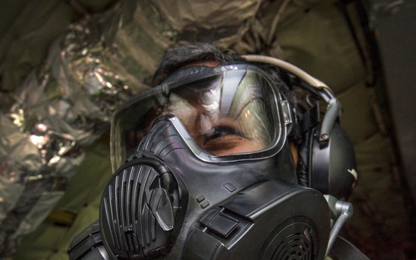 A person wearing a military-style gas mask or CBRN (Chemical, Biological, Radiological, Nuclear) protective mask is photographed in close-up inside what appears to be a military aircraft or vehicle. The individual is dressed in an olive/sage green flight suit or military uniform. The image appears to document military protective equipment use or training.