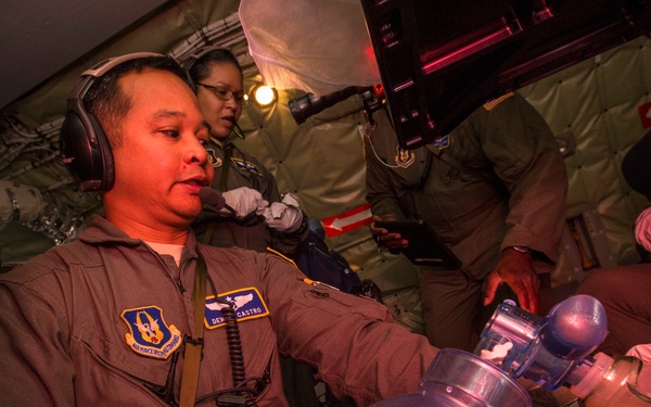 Military medical personnel, identified as 'Dennis Castro' based on the visible name tag, are performing an in-flight medical procedure aboard what appears to be a military transport aircraft. The primary figure is administering oxygen or ventilation using a bag-valve mask (BVM/Ambu bag) to a patient. Multiple crew members in flight suits are assisting or observing the procedure.