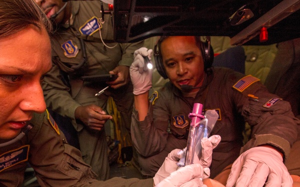 U.S. Air Force personnel in flight suits are conducting an in-flight medical training exercise aboard a military aircraft. Two primary personnel wearing latex gloves are practicing an airway management procedure on a medical training mannequin. A third person in the background appears to be observing or assisting with a penlight.