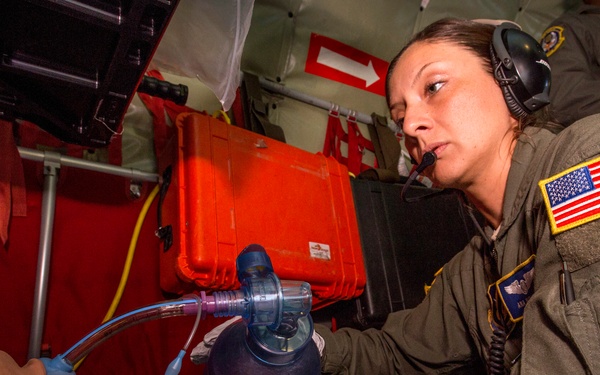 A U.S. military service member in an olive drab flight suit is performing a medical training exercise inside what appears to be a military aircraft. She is administering a bag-valve mask (BVM) to a medical training mannequin, simulating emergency airway management. The setting includes visible medical equipment cases and communication headsets, indicating an aeromedical training scenario.