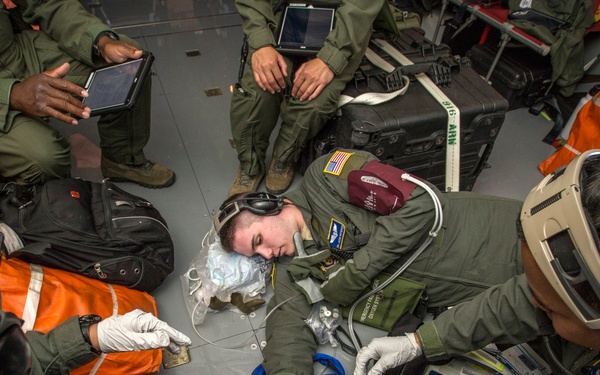 Military medical personnel in olive green flight suits are conducting what appears to be a medical training exercise or actual casualty care inside a military aircraft. A simulated or actual patient is lying on the floor surrounded by medics wearing gloves and specialized equipment including goggles. Multiple personnel are engaged with medical devices, IV lines, and monitoring equipment.