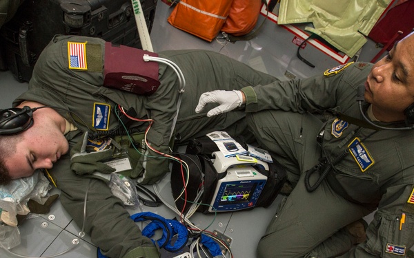 Two military personnel in olive green flight suits are conducting what appears to be a medical training exercise or actual casualty care aboard a military aircraft. One individual is lying on the floor in a simulated patient role while another, wearing white medical gloves and a flight helmet, attends to them using medical monitoring equipment. Multiple pieces of medical equipment, including a cardiac monitor/defibrillator, are deployed around them.