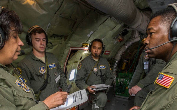 Four U.S. Air Force personnel in olive drab flight suits are gathered inside a military aircraft, appearing to conduct a briefing or evaluation. The individual on the left wears a 'Flight Examiner, 914 Aeromedical Evacuation Squadron' patch and is reviewing documents on a clipboard. All personnel are wearing Bose aviation headsets, suggesting active flight or pre-flight operations.
