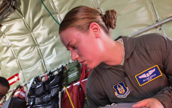 Two female U.S. Air Force personnel in olive flight suits are working inside what appears to be a military aircraft configured for aeromedical evacuation. The foreground subject, identified by her name tape as 'Jillian Moran,' is reviewing a binder while operating a medical monitor labeled 'PROPAQ.' A second airman works in the background among medical and tactical equipment.