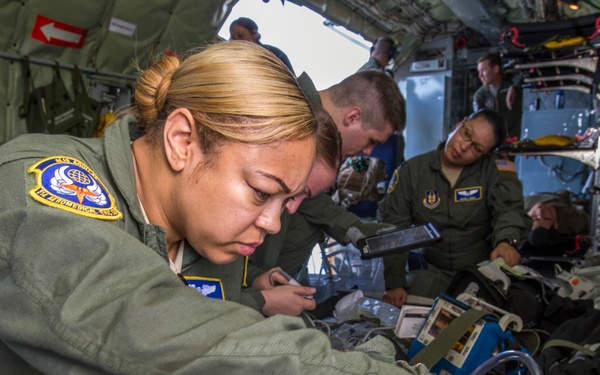 Multiple U.S. Air Force personnel in olive green flight suits are working inside what appears to be a military transport aircraft configured for aeromedical evacuation. The foreground shows a female airman closely examining or adjusting medical equipment, with a tablet device and medical gear visible nearby. Several other personnel are visible in the background performing similar tasks.