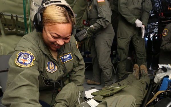 A smiling female U.S. Air Force personnel wearing an olive green flight suit is attending to a patient or mannequin on the floor of a military aircraft, likely during an aeromedical evacuation training exercise or mission. She is wearing aviation headphones with a boom microphone and has unit patches visible on her sleeve, including one reading 'Aeromedical Evac' with 'Alis Aquilae' text. Additional personnel in flight suits are visible in the background.