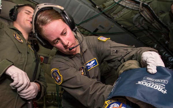 Military medical personnel in flight suits are conducting an aeromedical evacuation training exercise inside a military aircraft. A flight medic wearing latex gloves is attending to a patient lying on a litter, applying what appears to be a blood pressure cuff or medical device. Multiple personnel in olive drab flight suits with unit patches are present, suggesting a training or operational aeromedical scenario.