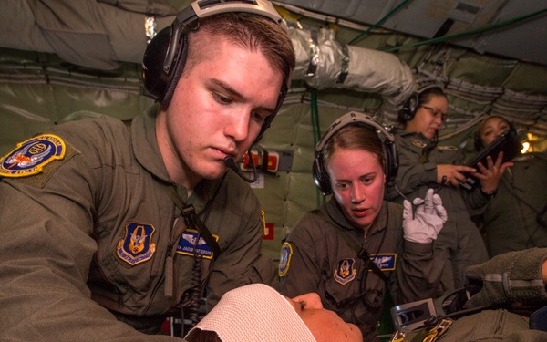 Military medical personnel, likely U.S. Air Force aeromedical evacuation crew members, are treating a patient aboard a military aircraft. The personnel are wearing olive drab flight suits with unit patches and aviation headsets, performing what appears to be in-flight medical care. At least four crew members are visible, with two actively engaged in patient treatment and others monitoring or documenting.