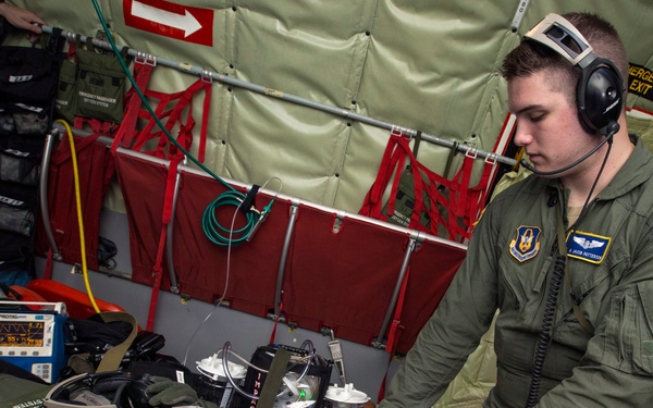A U.S. Air Force medical personnel wearing headset and latex gloves is attending to a patient lying on a stretcher inside a military aircraft. The scene appears to be an aeromedical evacuation exercise or actual medical transport, with the patient's head stabilized on a yellow cervical support pillow. Multiple medical monitoring devices and equipment are visible throughout the aircraft interior.