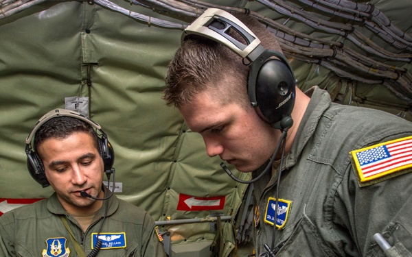 Two U.S. Air Force personnel in olive drab flight suits are reviewing information on a tablet device inside what appears to be a military transport aircraft. Both individuals are wearing aviation headsets with boom microphones, suggesting active communication during flight operations. The airman on the right displays a U.S. flag patch on his shoulder, consistent with standard military uniform regulations.