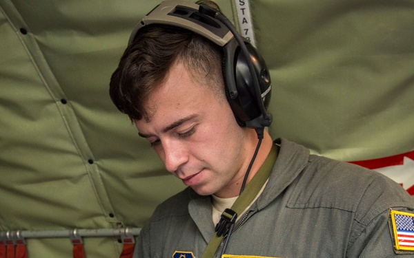 A U.S. Air Force airman identified as Stephen Karoly, rank A1C (Airman First Class), is reviewing documents in a binder while aboard a military aircraft. He is wearing a flight suit with a name tag, aviator wings badge, U.S. flag patch, and a unit patch, along with aviation headset communications equipment. The interior setting appears to be the cargo or crew area of a military transport aircraft.