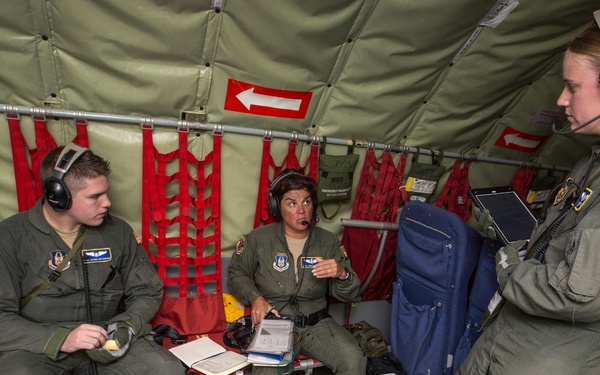 Three military personnel in olive green flight suits are gathered inside what appears to be a military transport aircraft cargo bay. They are engaged in a briefing or discussion, with two seated and one standing, all wearing communication headsets. The interior shows typical military aircraft features including red cargo netting, safety signage, and equipment storage.