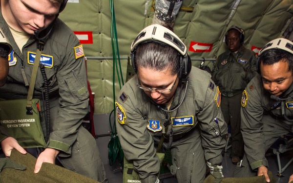 Military personnel in olive green flight suits are conducting what appears to be an aeromedical evacuation training exercise aboard a military aircraft. Several crew members wearing flight helmets with communications headsets are attending to a patient lying on a litter/stretcher covered with an olive green blanket. The setting appears to be the interior of a military transport aircraft configured for medical evacuation.