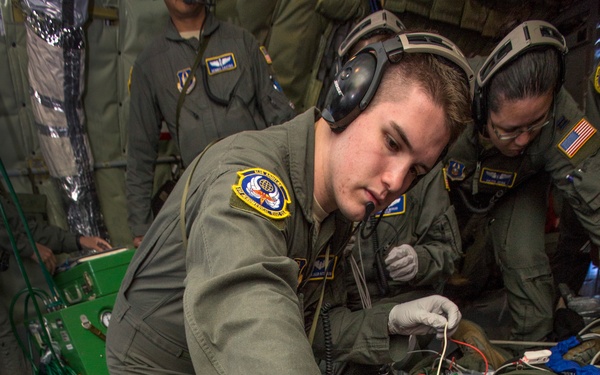 Military medical personnel in olive green flight suits are performing aeromedical evacuation procedures on a patient lying on the floor of what appears to be a military transport aircraft. At least three uniformed personnel are visible, with one wearing communication headsets and white latex gloves actively attending to the patient. The scene depicts an in-flight medical training exercise or actual casualty care operation.