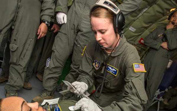 A military medical training or aeromedical evacuation exercise taking place inside what appears to be a military transport aircraft. A female service member wearing a flight suit and headset is performing a medical procedure on a patient lying on the floor, also in a flight suit. Several other uniformed personnel are observing in the background.