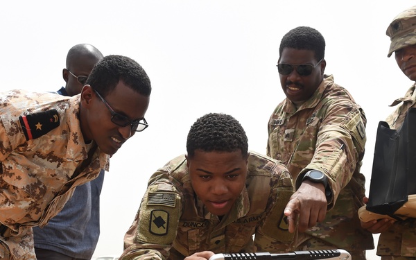Military personnel from what appears to be the U.S. Army and a partner nation's armed forces are gathered around a Panasonic Toughbook rugged laptop in an arid outdoor environment. The soldiers appear to be conducting a demonstration or training exercise involving the ruggedized computing equipment. The setting suggests a field operation or joint military exercise in a desert or semi-arid region.