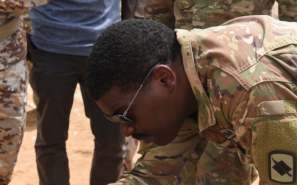 A U.S. Army soldier in OCP (Operational Camouflage Pattern) uniform is leaning over and operating a ruggedized Panasonic Toughbook laptop in an outdoor, arid environment. The soldier wears glasses and a unit patch visible on the left sleeve. Several other individuals in mixed military attire are present in the background, suggesting a multinational training or operational context.