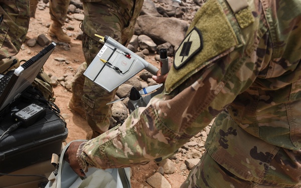 Military personnel in MultiCam/OCP camouflage uniforms are handling tactical equipment in an arid, rocky outdoor environment. A soldier is retrieving or stowing what appears to be a small fixed-wing UAV (unmanned aerial vehicle) from a carrying bag, while ruggedized computing equipment and cases are stacked nearby. The unit patch visible on the right soldier's shoulder appears to be a lightning bolt insignia.