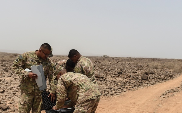 Three military personnel in MultiCam/OCP camouflage uniforms are gathered around a tan hard-sided equipment case on a dirt road in an arid, rocky desert environment. One soldier holds a document or folder while the others examine the open case, which bears a biohazard symbol on its side. The landscape is barren with scattered rocks, sparse vegetation, and a hazy sky consistent with a hot, dry climate.