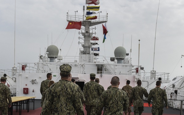 A group of military personnel in camouflage uniforms walk across the deck of a white naval vessel toward the ship's superstructure. A Chinese national flag is prominently displayed on the mast alongside other signal flags, suggesting this may be a Chinese naval or hospital ship. Red cross symbols are visible on equipment covers on both sides of the deck, indicating this is likely a medical or hospital ship.