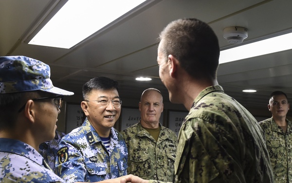 The image depicts a formal military meeting or exchange between personnel wearing Chinese People's Liberation Army Navy (PLAN) uniforms and what appear to be U.S. Navy personnel in digital camouflage uniforms. A senior PLAN officer is shaking hands with a U.S. Navy servicemember in what appears to be a shipboard conference or briefing room. The setting suggests an official bilateral military engagement or port visit.