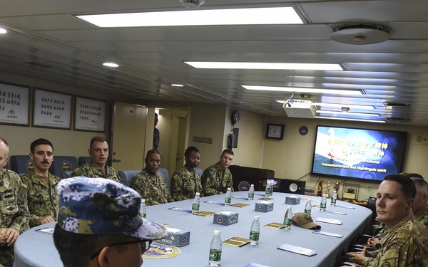 A formal military meeting is taking place around a large oval conference table aboard what appears to be a naval vessel. Personnel in two distinct uniforms are present: Chinese People's Liberation Army Navy (PLAN) digital camouflage uniforms and U.S. Navy/military uniforms in NWU (Navy Working Uniform) and OCP patterns. A presentation screen displaying Chinese and English text referencing 'Humanitarianism and Nightingale spirit' is visible in the background.