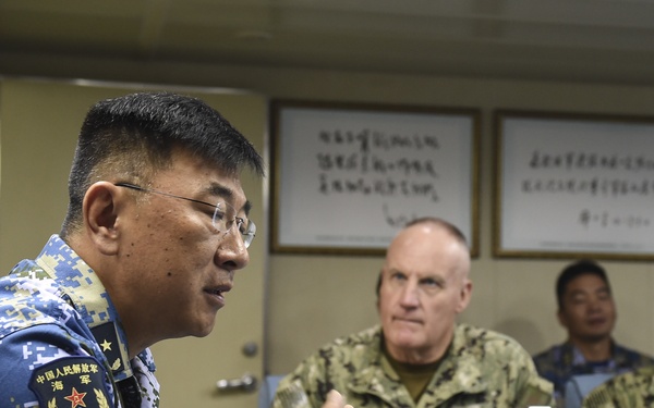 A military meeting or briefing is taking place between personnel from the Chinese People's Liberation Army Navy (PLAN) and what appear to be United States Navy officers. The PLAN officer on the left, wearing blue digital camouflage with visible Chinese military insignia, appears to be speaking or presenting. The setting appears to be aboard a naval vessel, indicated by the institutional interior and framed documents on the wall.