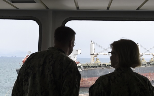 Two individuals in military camouflage uniforms are observed from behind, standing inside a vessel or building and looking through large windows at a large bulk carrier cargo ship docked nearby. The cargo ship features a distinctive red hull with visible loading cranes and appears to be moored at a port facility. The scene suggests a military observation or inspection activity at a maritime location.